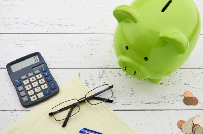 A piggy bank, calculator, pen, glasses, and money arranged on a white wooden table
